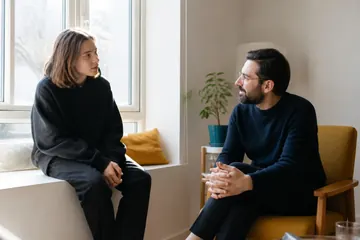 Omuz hizasında saçlı bir genç, pencere kenarındaki oturma yerinde sakallı bir psikologla güneşli bir odada konuşuyor